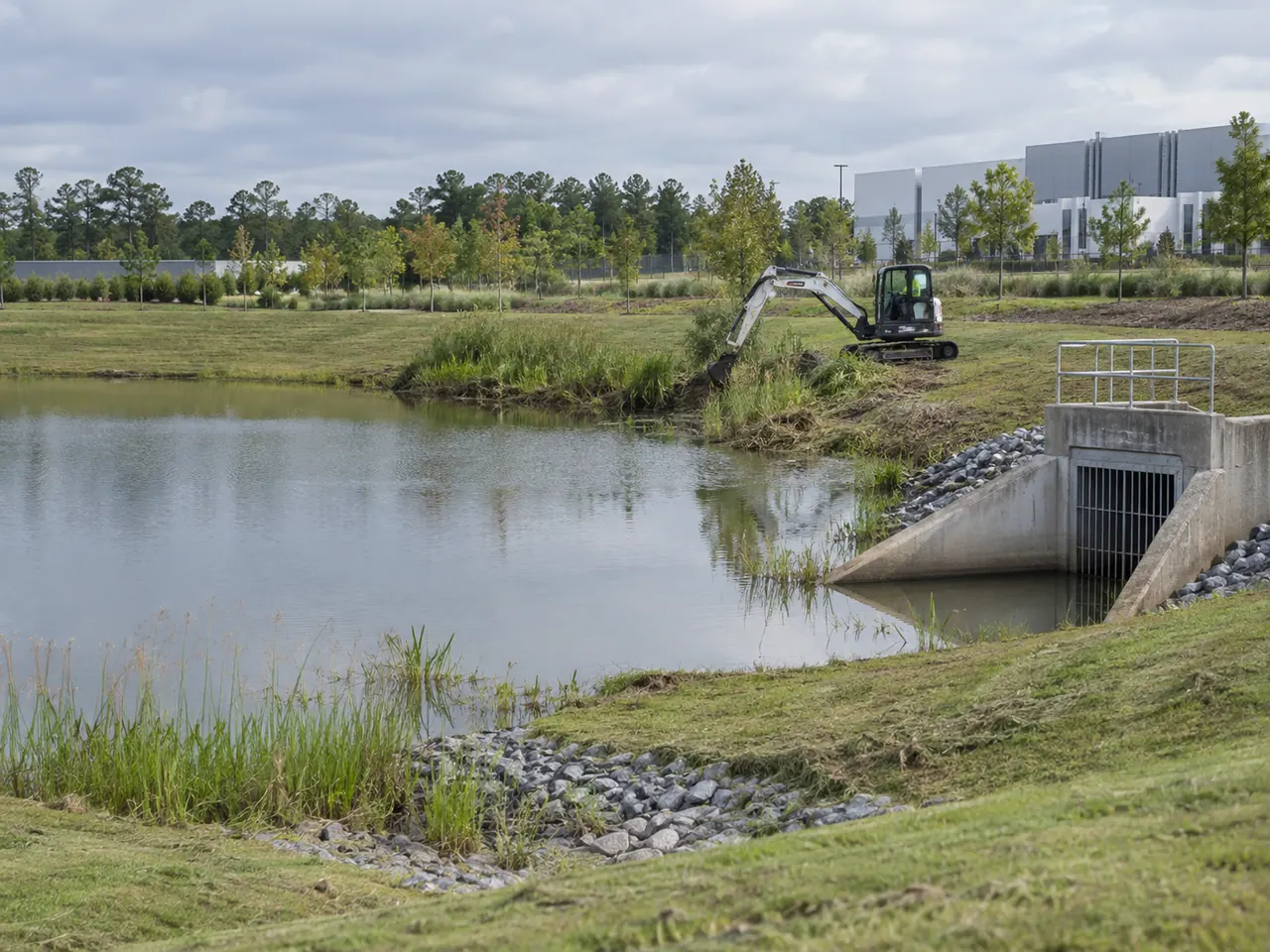 Compact excavator maintaining vegetation beside a commercial stormwater pond and outlet structure